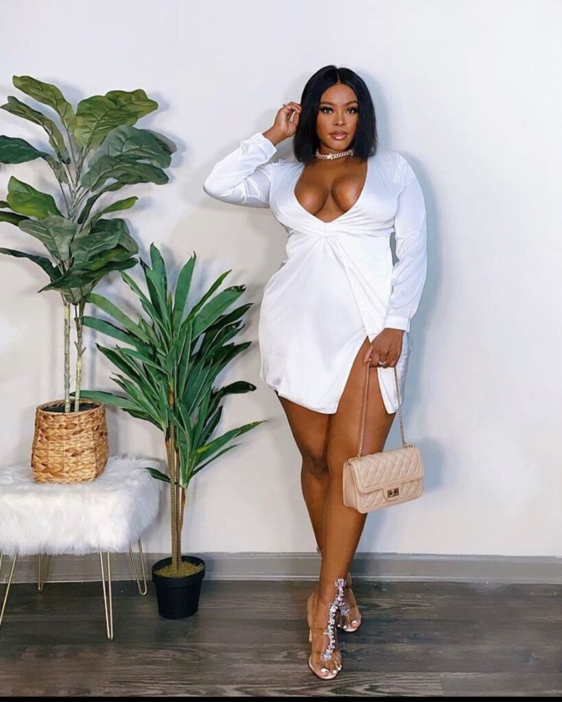 In an indoor setting, a woman dons a white dress and heels, exuding style and grace. The backdrop includes a houseplant and a flowerpot, adding a touch of natural beauty.
