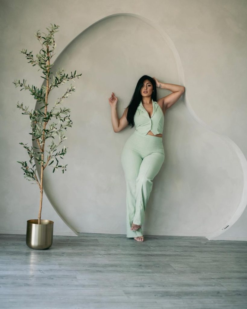 Woman in mint green jumpsuit leaning against wall, indoor setting with houseplant.