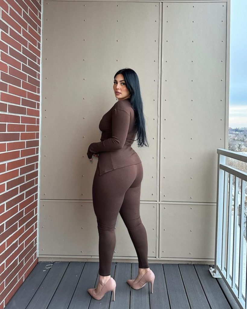 Fashionable woman in brown bodysuit and heels standing on balcony, exuding elegance against building background.