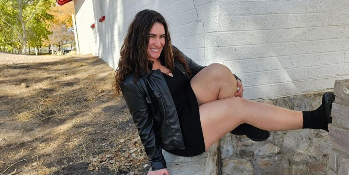 An elegant woman with long hair sits on a rock wall, donning a leather jacket and black boots. Her radiant smile complements her fashionable attire.