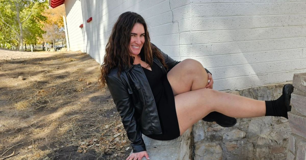 An elegant woman with long hair sits on a rock wall, donning a leather jacket and black boots. Her radiant smile complements her fashionable attire.