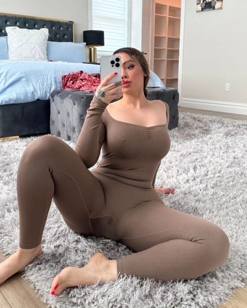 Woman in brown bodysuit taking a selfie indoors on a bed, wearing pantyhose. Focus on lower body and upper torso.