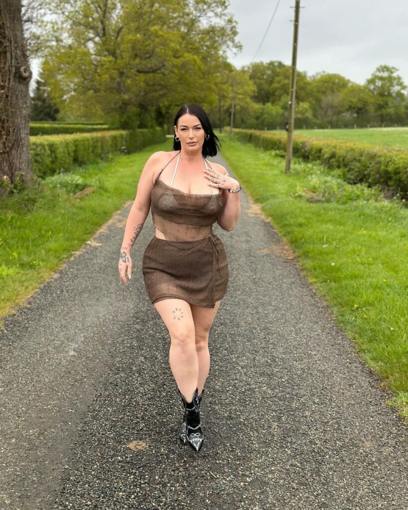 "A woman wearing a brown dress strolls along a road, with a scenic view of grass and trees in the background."
