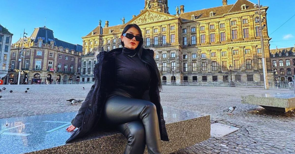 Stylish woman in black leather pants and coat sitting on bench by fountain in city setting.
