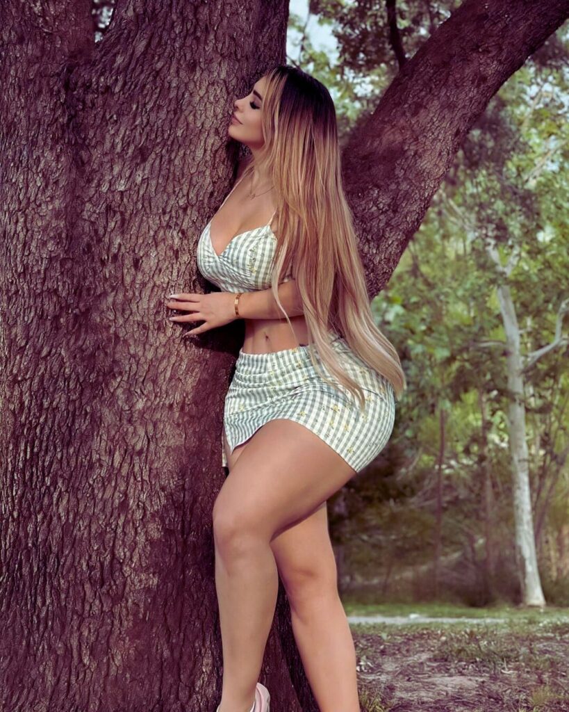 A fashionable woman with long hair leans against a tree, posing for a photo shoot in a short dress.