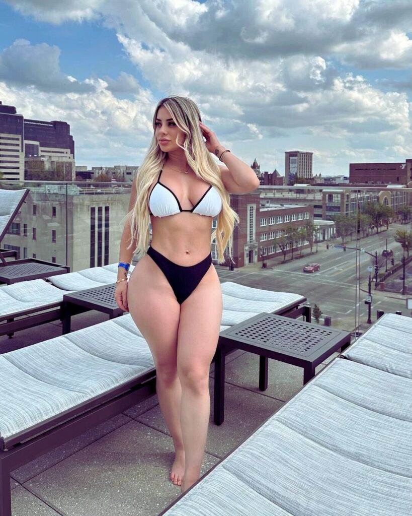 A woman in a bikini posing on a rooftop with sky and clouds in the background.