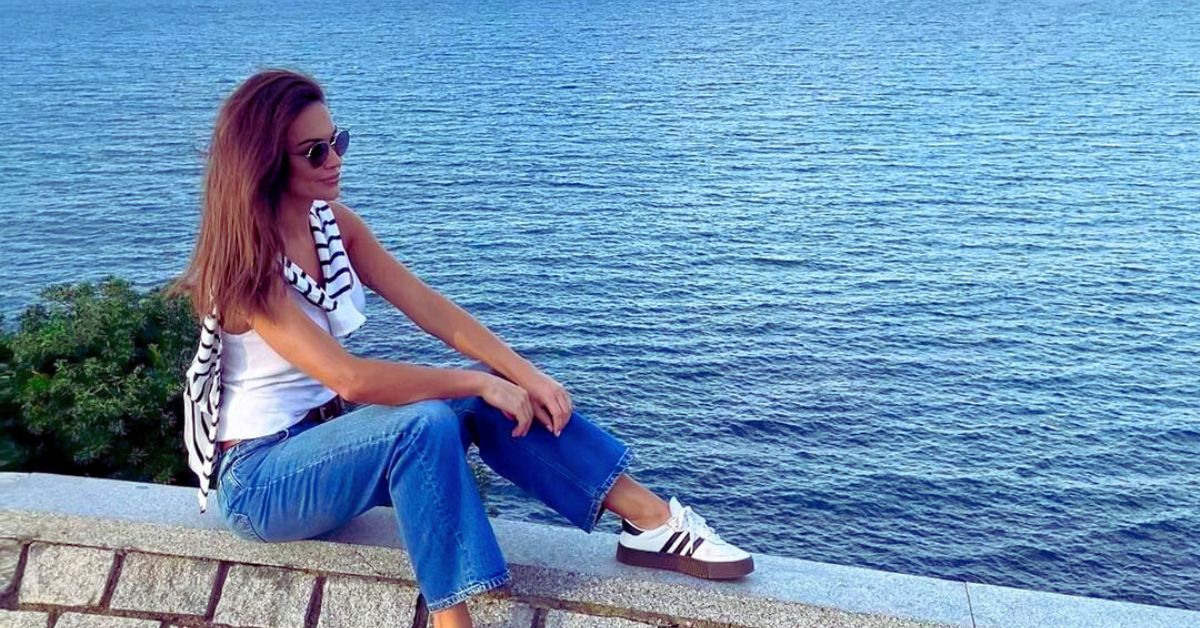 Serene woman in jeans sitting on a ledge by the ocean, enjoying the scenic view of a calm lake on a summer day.