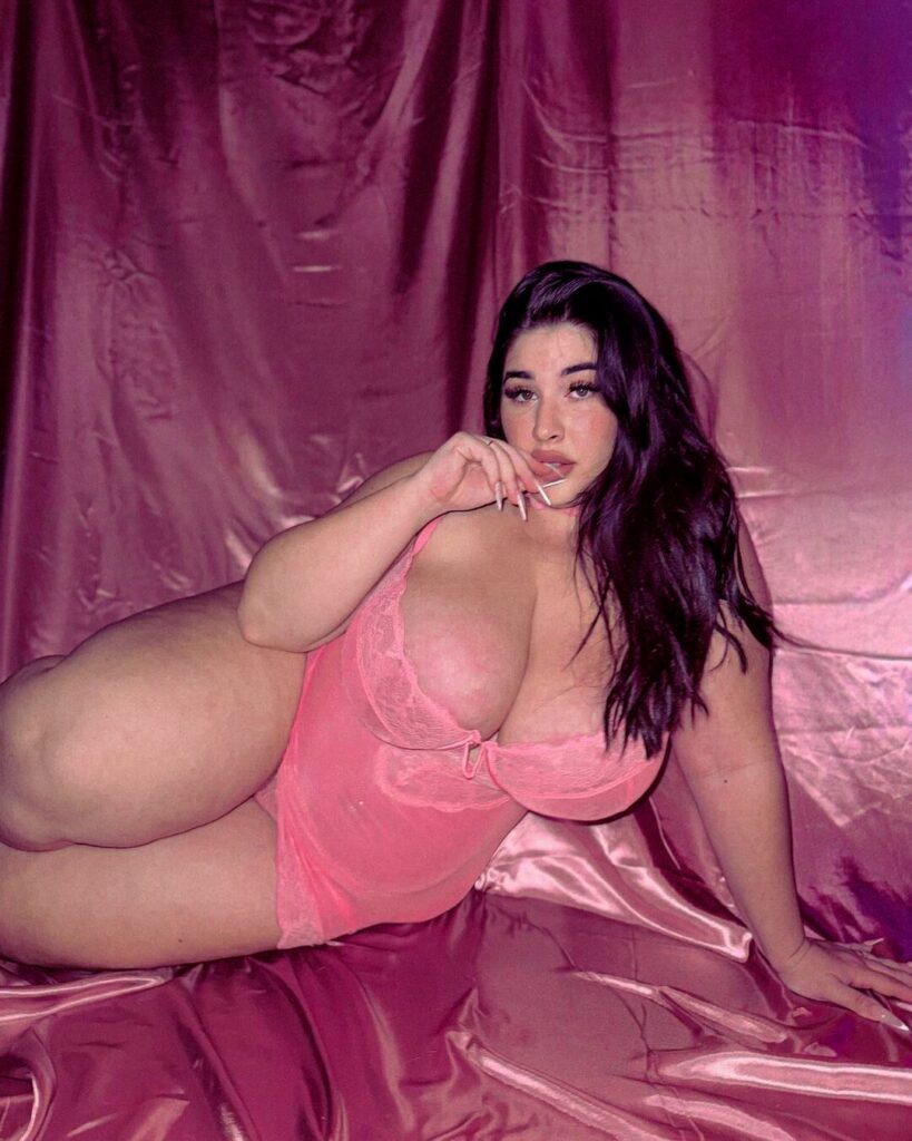 Fashion model in pink lingerie, lying on a pink sheet for a photo shoot, showcasing clothing and style.